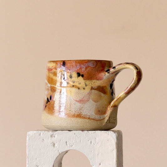 A handcrafted stoneware mug with a painterly design, featuring pink, yellow, tan, peach and pearly blue tones, and black dots, displayed on a white stand against a beige background.
